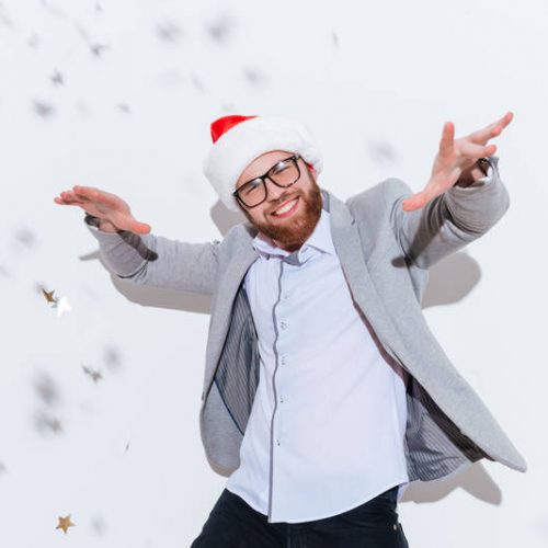 Cheerful bearded young man in glasses and santa claus hat smiling and dancing over white background